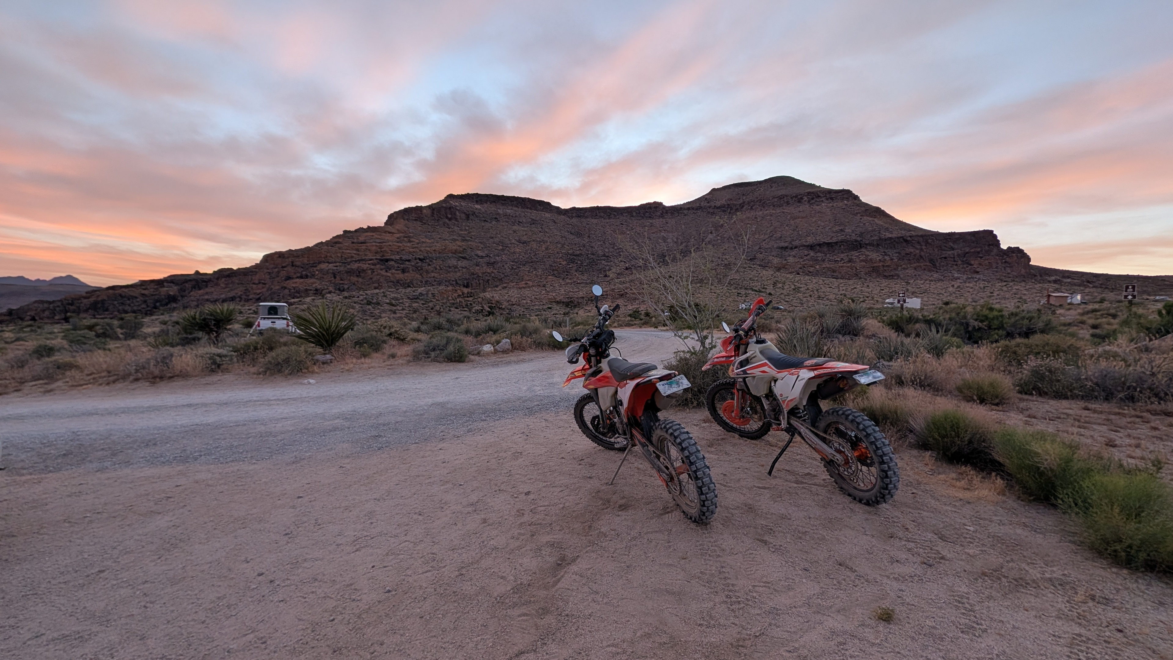 Two red and white off-road dirt bikes parked on a dirt road in a desert landscape with a large rock formation and a pink and orange sunset sky in the background.